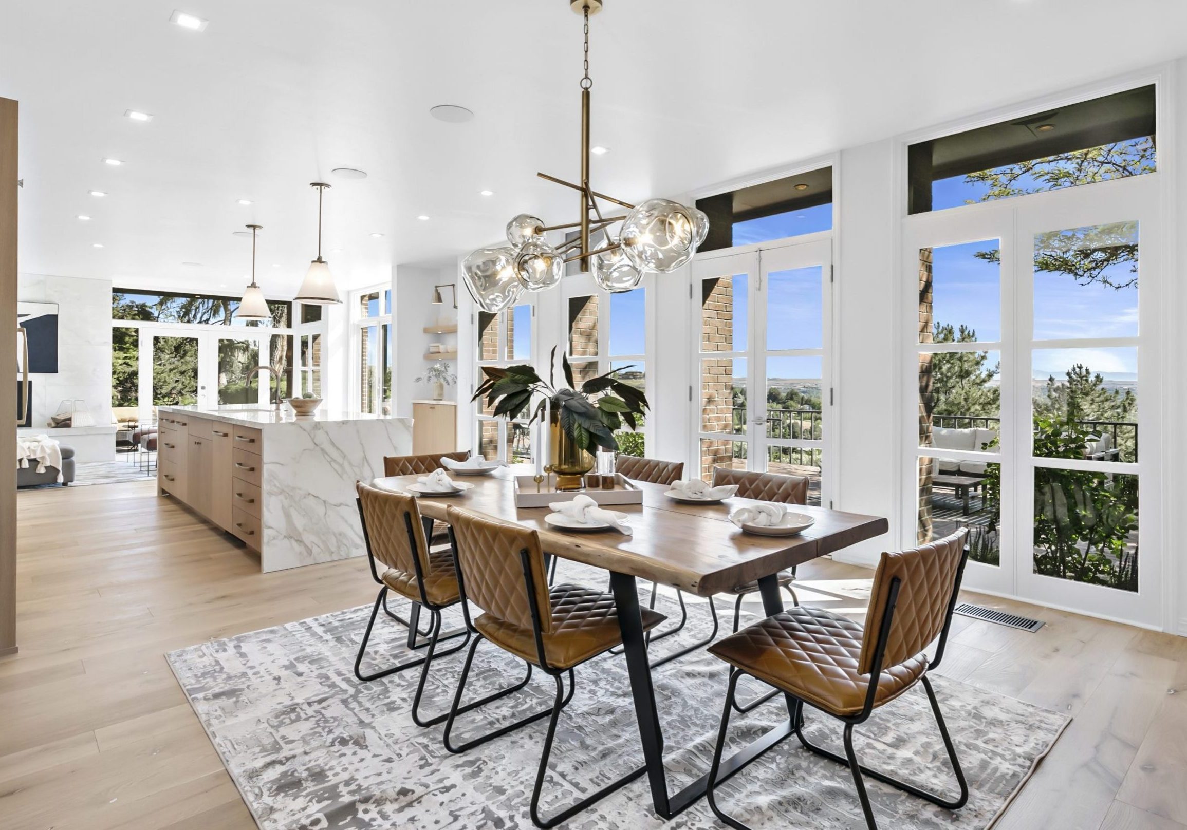 Stunning luxury interior view of dining space into the modern kitchen. Bright white wall of windows and french doors that lead to the back patio and overlook of the Treasure Valley