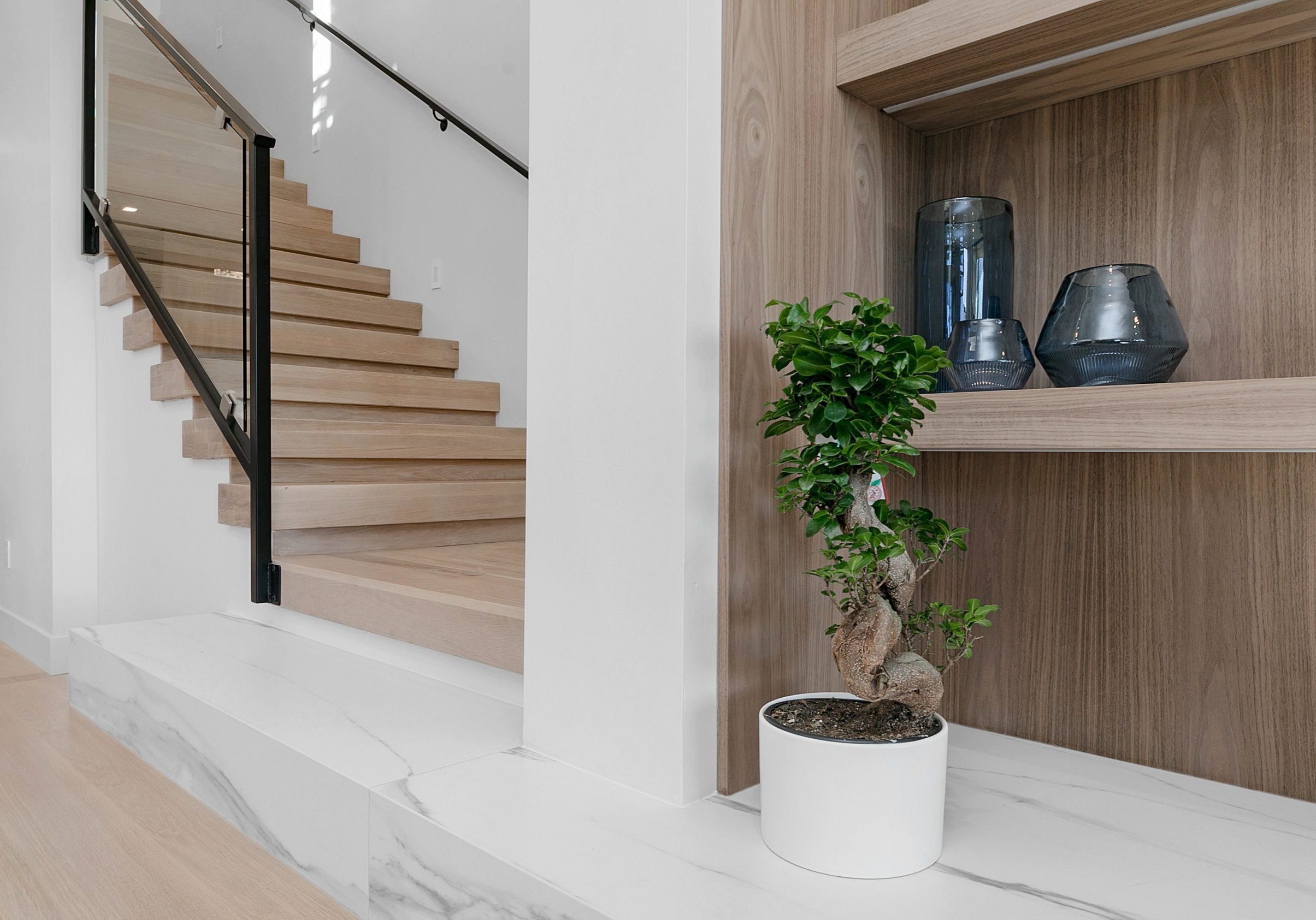 Detail view of contemporary, light wood staircase with black metal and glass banisters. A marble step leads across with built in shelving and plant